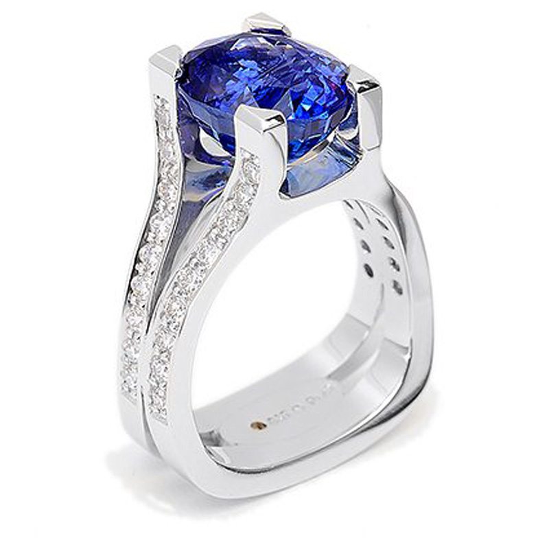 A signature Paragon-shaped engagement ring featuring a massive, cushion-cut blue sapphire held by four heavy platinum prongs. The band's "Shape of the Finger" design is engineered for comfort and to prevent spinning, with its split-shank sides adorned with recessed pavé diamonds.